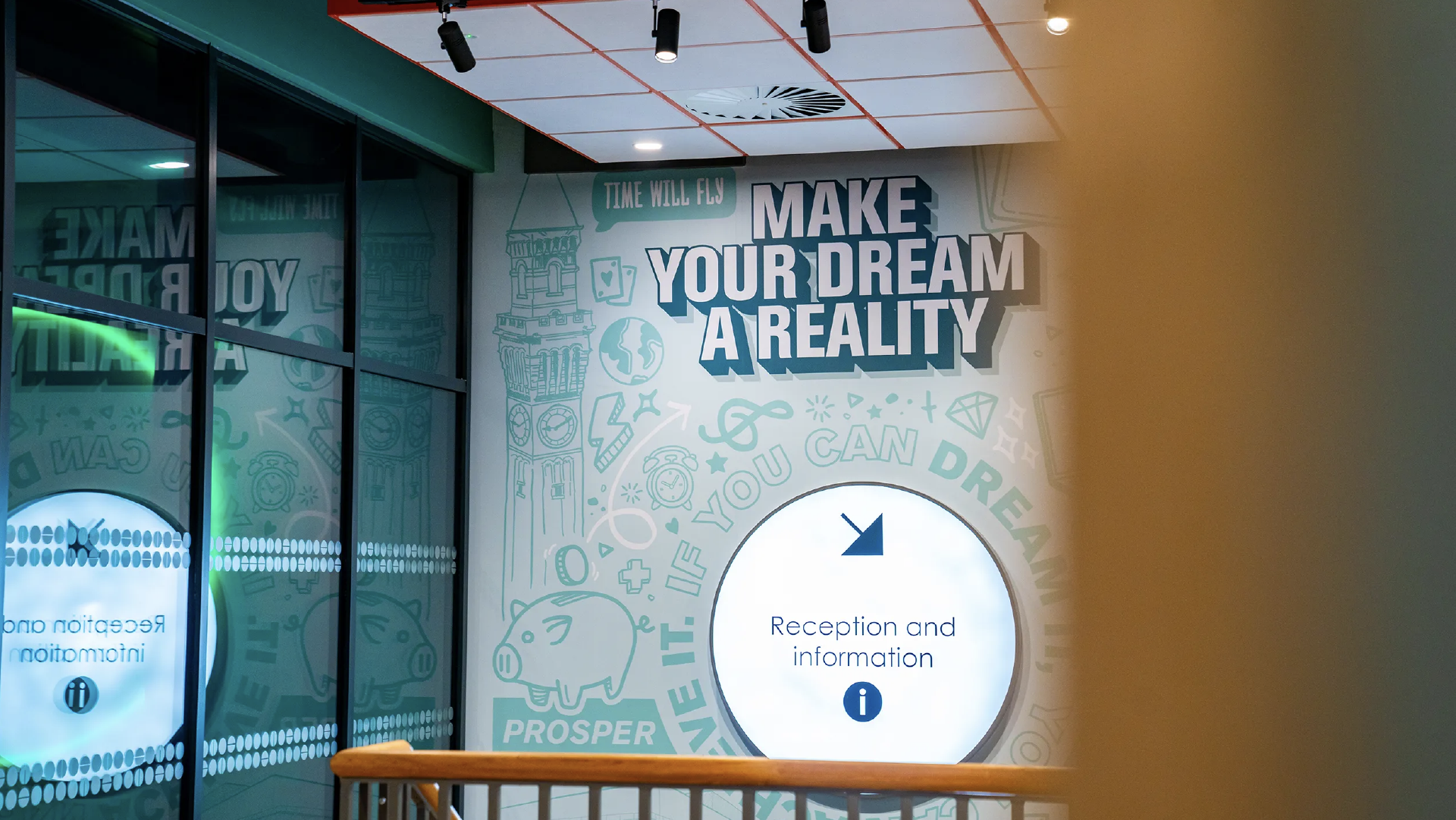 Reception wall reading 'Make your dream a reality' over teal line illustrations, beside a circular reception and information sign
